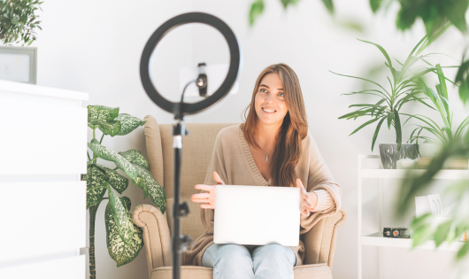 Woman sitting on chair looking towards tripod with phone as she is smiling at camera